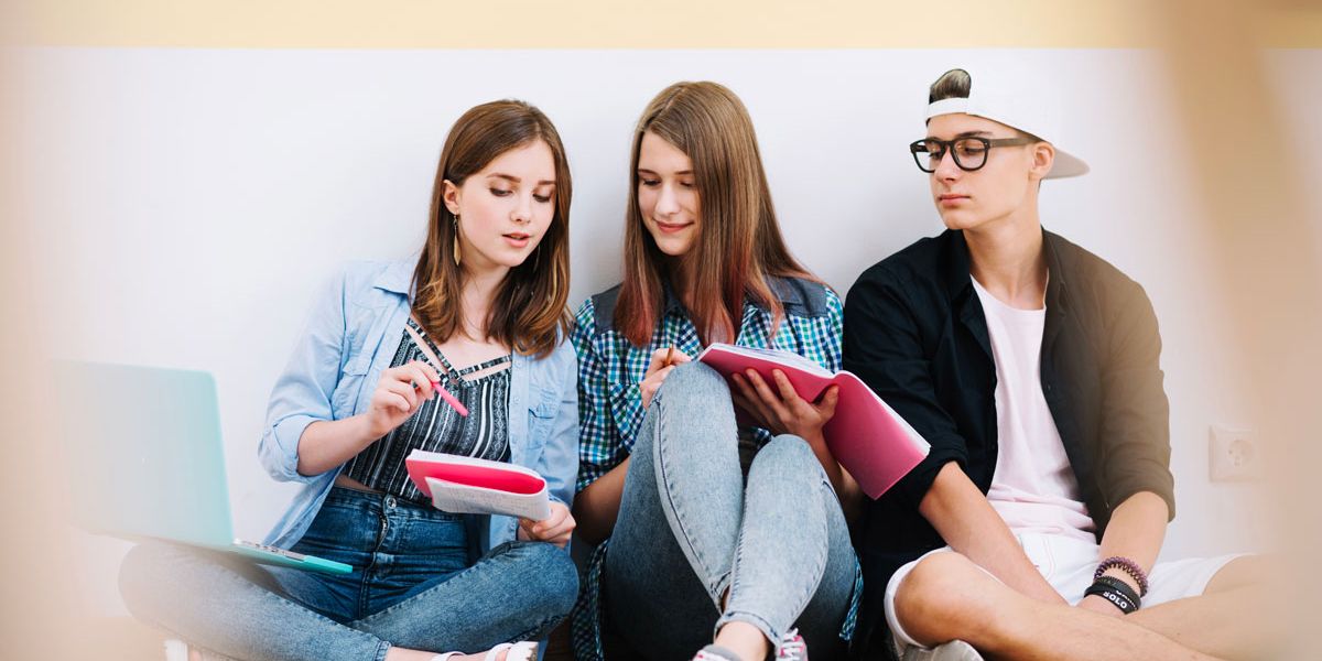 Three young people sitting on the floor, engaged in writing notes in their notebooks.